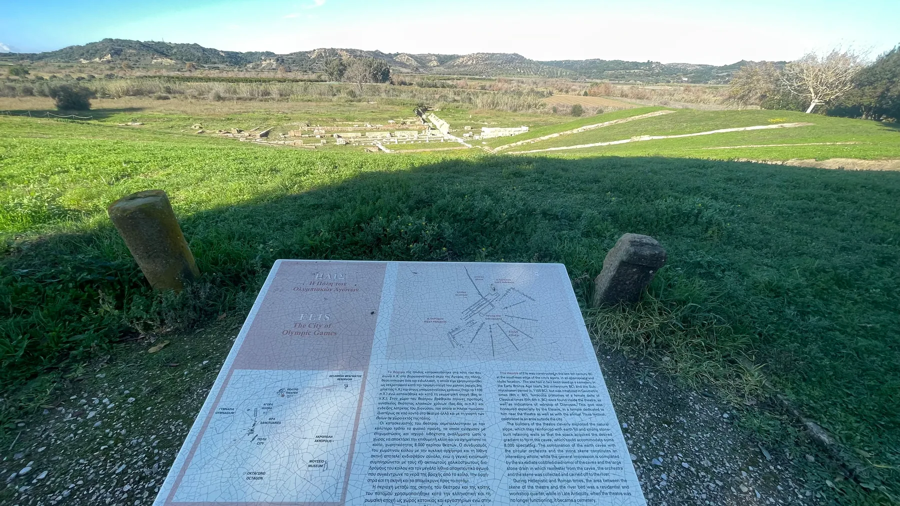 The bilingual info plaque at Ancient Elis, reading 'ELIS: The City of Olympic Games', ruins visible below