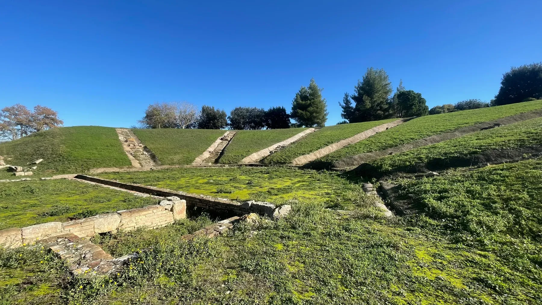 Grass-covered banked terraces and stone ruins at Ancient Elis