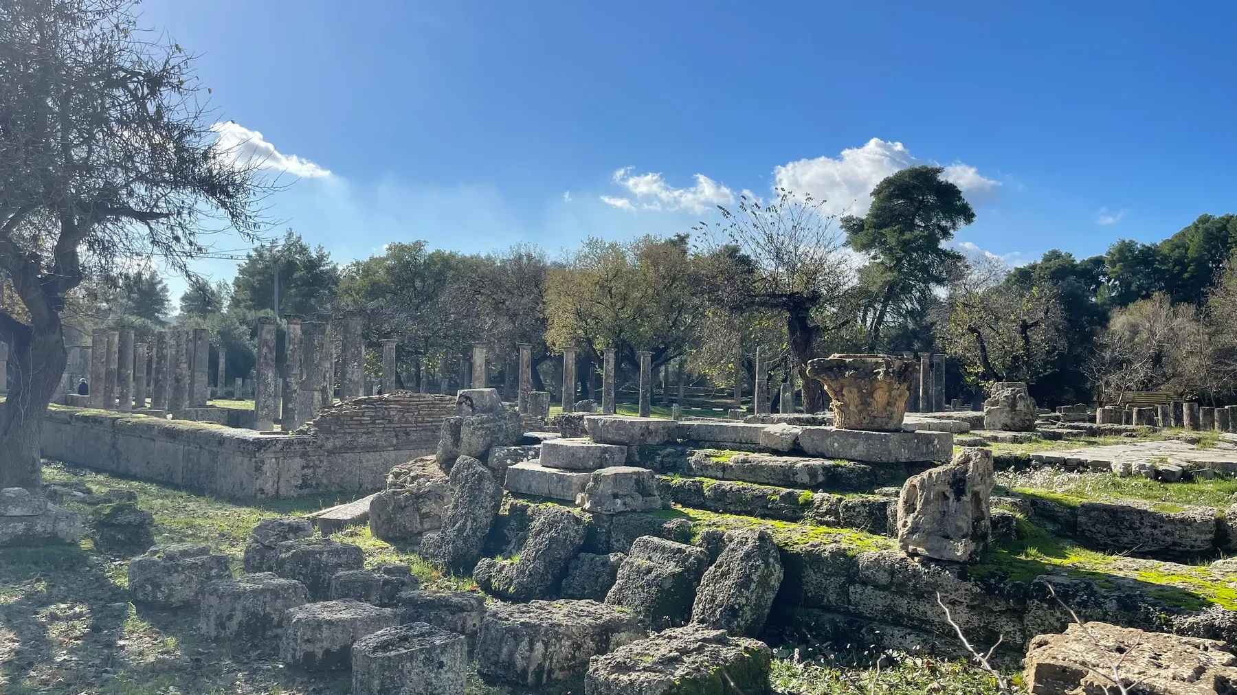 A carved Corinthian capital resting among fallen stone blocks at Olympia, columns beyond
